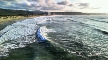 Güzel dalgalar zarif bir şekilde kumlu bir plaja yuvarlanıyor. Uzak binalar, çarpıcı bir günbatımı gökyüzünün altında görülebilir.