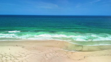Sakin ve sakin bir sahil manzarası güzel turkuaz suları, yumuşak dalgaları ve berrak bir gökyüzünün altında yumuşak kumları sergiliyor.