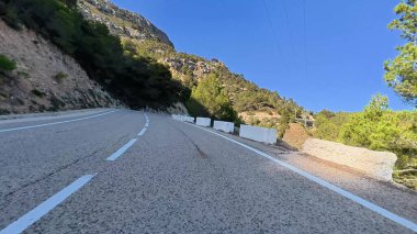 Uzun ağaçlar ve açık mavi gökyüzü ile güzel bir şekilde çizilmiş kıvrımlı bir dağ yolunun nefes kesici manzarası.