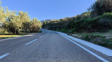 Açık mavi gökyüzünün altında, zeytin ağaçları ve kayalık tepelerle çevrili dolambaçlı bir yol manzarası..