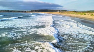 Okyanus dalgalarının büyüleyici ve büyüleyici görüntüsü açık mavi gökyüzünün altındaki kumlu bir sahile çarpıyor.