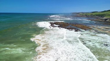 Açık mavi gökyüzünün altında dalgalar, kayalar ve yeşilliklerle kıyı şeridinin çarpıcı hava manzarasının tadını çıkarın.