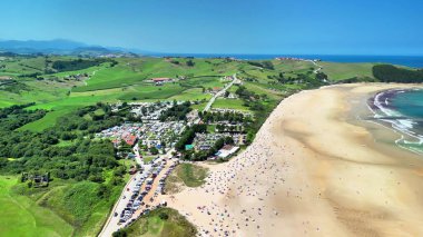 Huşu uyandıran bir hava görüntüsü, açık mavi gökyüzünün altında yuvarlanan yeşil tepelerin yanında el değmemiş bir sahili yakalar.