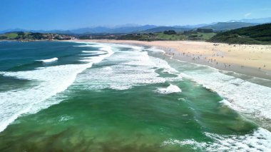 Yumuşak dalgaların, yemyeşil tepelerin ve sıcak güneşin tadını çıkaran mutlu insanların olduğu güzel ve pitoresk bir sahil sahnesi.