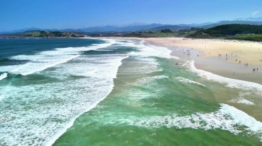 Hava görüntüsü dalgaların kumlu bir sahile çarptığını gösteriyor. Neşeli insanlar günlerini dolu dolu geçiriyorlar.