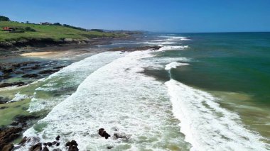 Güzel bir kıyı şeridinin çarpıcı hava manzarası, açık mavi gökyüzünün altında yemyeşil tepelere çarpan dalgalar.