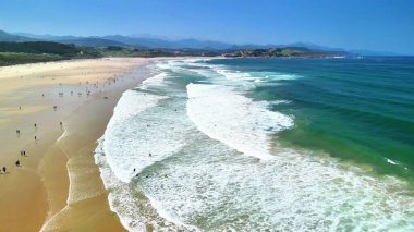 Güzel bir sahil berrak suları, dalgaları, sörfçüleri ve neşeli kumsalcıları gösterir. Güneşin tadını çıkarırlar.