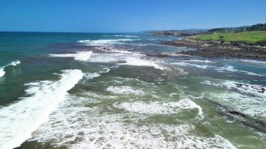 Bir hava görüntüsü okyanus dalgalarının engebeli bir kıyı şeridine çarpıp açık mavi gökyüzünün altında güzelliği yaratmasını sağlar.