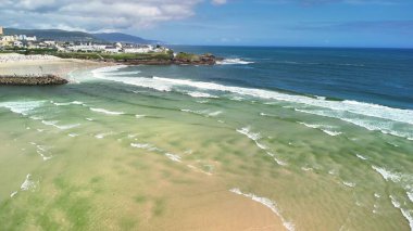 Hava görüntüsü, mavi gökyüzünün altındaki kumlu sahillere çarpan canlı dalgaları yakalıyor. Macera ve sükuneti çağrıştırıyor.