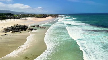 Sakin bir plajın sersemletici hava manzarası yumuşak dalgaların kumlu kıyıya hafifçe vuruşunu gösteriyor.