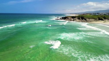 Çarpıcı bir hava manzarası, berrak mavi gökyüzünün altında canlı turkuaz suları ve kayalık kıyı şeridini vurguluyor