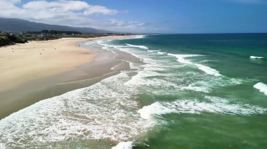 Hava görüntüsü, berrak mavi gökyüzünün altında sakin sahil dalgaları gösteriyor. Rahatlamak ve günlük stresten kaçmak için ideal.