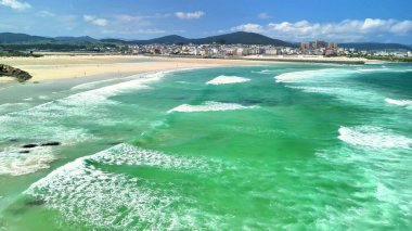 Sakin bir plajın güzelliğini gösteren, turkuaz suları ve çarpıcı bir şehrin siluetini gösteren bir hava fotoğrafı.