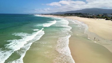 Bu, yumuşak, yuvarlanan dalgalar ve açık mavi gökyüzü içeren sakin bir plajın çarpıcı bir görüntüsü.