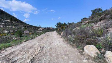 Resmedilmeye değer toprak bir yol, berrak mavi gökyüzünün altındaki yeşil tepeler ve çiçek açan bitkiler arasında dolanıyor.