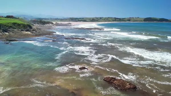 Büyüleyici kıyı şeridi manzarasında yumuşak dalgalar, kayalık oluşumlar ve yakınlardaki yemyeşil tepeler yer alıyor.
