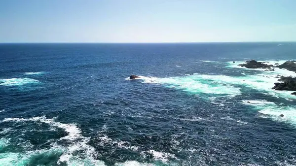 Açık mavi gökyüzünün altındaki kayalık bir sahile çarpan okyanus dalgalarının şaşırtıcı güzellikteki manzarasının tadını çıkar.