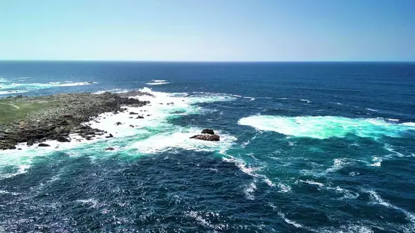 Sakin mavi gökyüzünün altındaki kayalık kıyıların ve canlı turkuaz dalgaların nefes kesici ve çarpıcı hava manzarası.