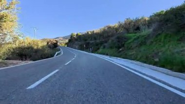 Açık mavi gökyüzünün altında, zeytin ağaçları ve kayalık tepelerle çevrili dolambaçlı bir yol manzarası..