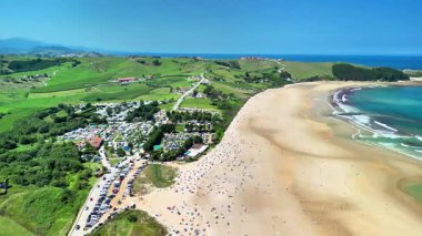 Huşu uyandıran bir hava görüntüsü, açık mavi gökyüzünün altında yuvarlanan yeşil tepelerin yanında el değmemiş bir sahili yakalar.