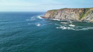 Açık mavi gökyüzünün altındaki dalgalar ve uçurumlarla kıyı manzarasını nefes kesici bir şekilde gözlemleyin.