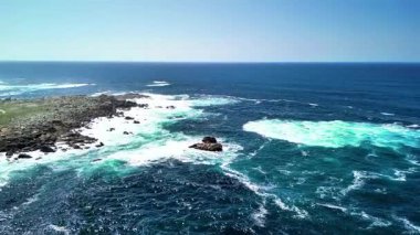 Sakin mavi gökyüzünün altındaki kayalık kıyıların ve canlı turkuaz dalgaların nefes kesici ve çarpıcı hava manzarası.