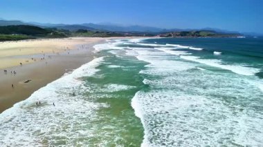 Çarpıcı bir hava manzarası dalgaların nazikçe çarptığı ve güneşlenenlerin mükemmel günün tadını çıkardığı canlı bir plajı sergiliyor.