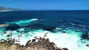 Çarpıcı kıyı manzarası dalgaların, berrak mavi gökyüzünün altındaki engebeli kayalara güçlü bir şekilde çarptığını gösteriyor.