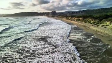 Yumuşak okyanus dalgalarının büyüleyici görüntüsü yukarıdaki bulutlu gökyüzünün altındaki kumlu kumsala yavaşça yuvarlanıyor.
