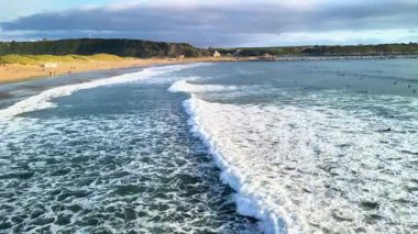 Kıyıya doğru yuvarlanan okyanus dalgalarının güzel manzarası. Sörfçüler bulutlu bir gökyüzünün altında suyun tadını çıkarıyorlar..