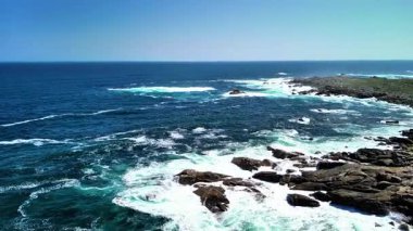 Okyanus dalgalarının berrak mavi gökyüzünün altında kayalık kıyılara çarpışına tanık olun.
