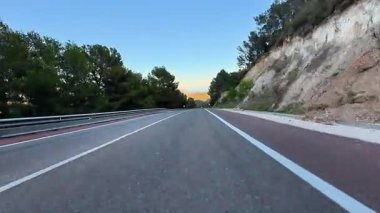 Canlı günbatımı gökyüzü, bereketli ağaçlar ve kayalık tepelerle çevrili dolambaçlı bir yola güzelce uzanıyor.