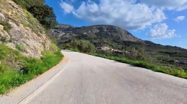 Dolambaçlı bir yol, yemyeşil ve yüksek dağların arasında bulutlarla süslenmiş parlak mavi bir gökyüzünün altında dolanıyor.