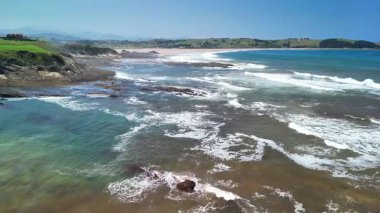 Büyüleyici kıyı şeridi manzarasında yumuşak dalgalar, kayalık oluşumlar ve yakınlardaki yemyeşil tepeler yer alıyor.