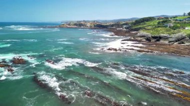 Sersemletici hava görüntülerinde sakin sahil şeridi, canlı mavi su ve parlak güneşli bir günde kayalık kıyılar sergileniyor.