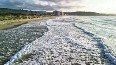 Sahile vuran güzel sahil dalgaları, gün batımında arka planda binalar ve tepeler.