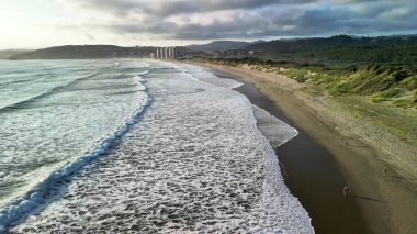 Şaşırtıcı derecede güzel bir plaj sahnesi dalgaları yakalıyor kumlu kıyıya yuvarlanan, canlı bir şehir siluetine yakın.