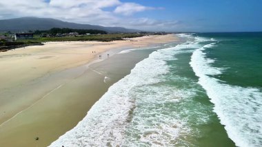 Sersemletici hava manzarası sakin bir plajı gösteriyor. Dalgalar açık mavi gökyüzünün altında kıyıya vuruyor.