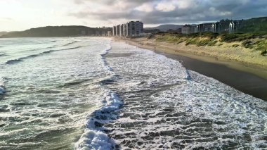 Çarpıcı bir kıyı manzarası sahile vuran dalgalar, modern binalar ve güzel bir günbatımı barındırır.
