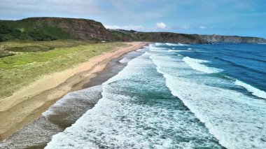 Göz kamaştırıcı bir hava manzarası sakin bir sahili gösteriyor. Dalgalar sahili öpüyor ve güzel yeşil kayalıkları gözler önüne seriyor.