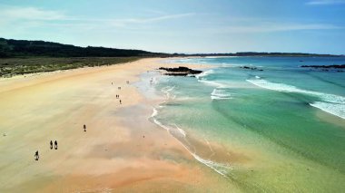 İnsanların güneşin altında harika vakit geçirdiği ve dalgaların tadını çıkardığı çarpıcı bir sahili gösteren bir hava manzarası.