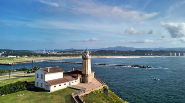 Tarihi bir deniz feneri, canlı bir rıhtım ve berrak mavi gökyüzünün altındaki dağlardan oluşan güzel bir kıyı manzarası.