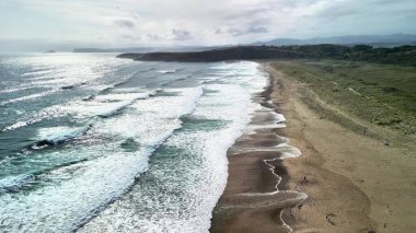 Sakin ve sakin bir sahil manzarası. Dalgalar bulutlu bir gökyüzünün altında kumlu kıyı şeridine yavaşça çarpıyor.