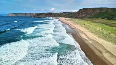 Uçurumlar ve yemyeşil yeşilliklerle çevrili kumlu bir plaja çarpan dalgaların nefes kesici görüntüsü..