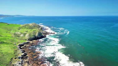 Bereketli yeşillik ve güçlü çarpan dalgalarla karakterize edilmiş canlı bir kıyı şeridini gösteren nefes kesici bir hava manzarası.