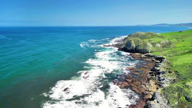 Arkasında yemyeşil tepeler bulunan kayalık kıyılara çarpan dalgaların çarpıcı kıyı manzarası