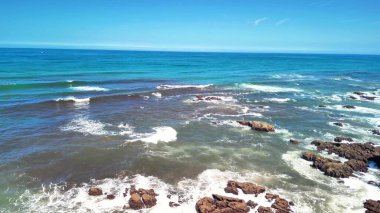 Parlak güneş ışığı ve açık mavi gökyüzü altında dalgaların engebeli kayalara çarptığı çarpıcı bir sahil şeridi.