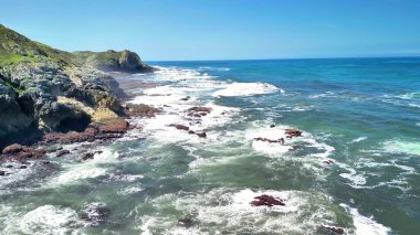 Görkemli okyanus dalgalarının açık mavi gökyüzünün altında kayalık kıyılara çarptığını gösteren nefes kesici bir sahne.