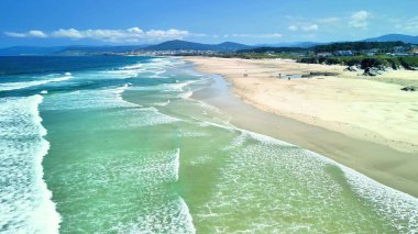 Bir hava fotoğrafı, kıyı şeridi boyunca berrak turkuaz suları ve yumuşak altın kumları olan sakin bir sahili yakalar.