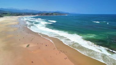 Çarpıcı bir sahil manzarası güneşlenenleri mutlu bir şekilde yakalıyor sıcak kumun ve berrak mavi suların tadını çıkarıyor.